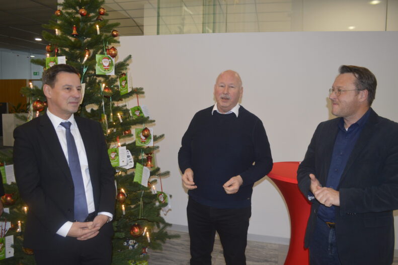 Drei Männer stehen vor zwei festlich geschmückten Weihnachtsbäumen mit Wunschkarten; sie unterhalten sich in freundlicher Atmosphäre, während die Bäume mit Kugeln und Lichtern im Hintergrund leuchten.